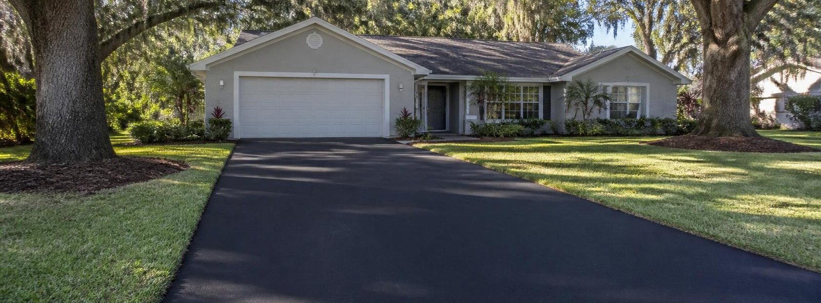 Freshly paved black asphalt driveway running to a gray Florida ranch home beneath moss-draped live oaks